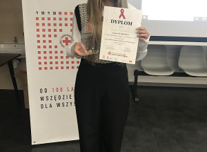 Oliwia Miętkiewicz uczennica klasy drugiej. Dziewczyna stoi na tle banera Polskiego Czerwonego Krzyża. W rękach trzyma dyplom za zajęcie 4 miejsca w konkursie oraz szklaną statuetkę.
